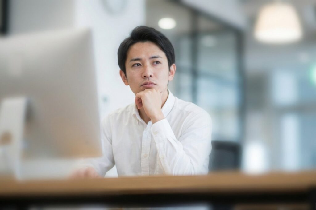 オフィスで顎に手を当てて座るビジネスパーソンとデスクトップPC。業務の停滞や迷いを一次予防の面談で整理する。
